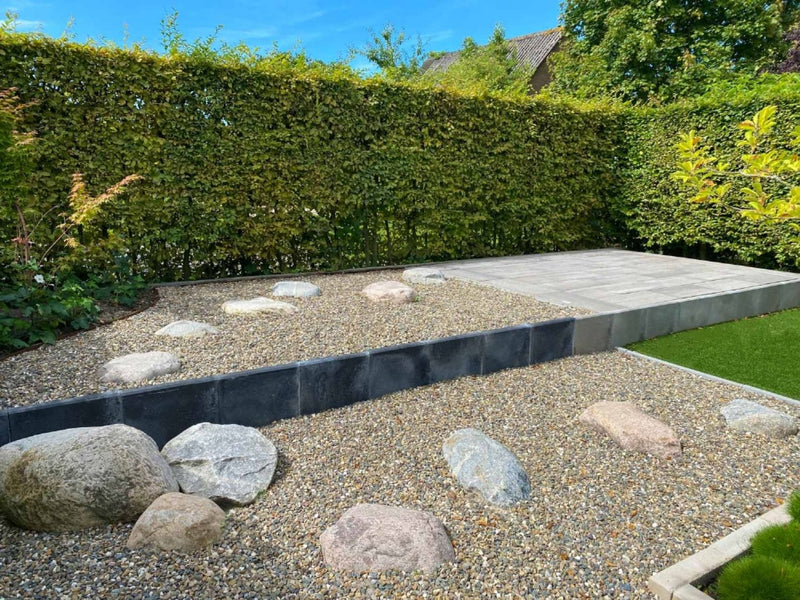 Een moderne tuin met een minimalistisch ontwerp bevat een grindgedeelte dat is versierd met grote, gladde stenen. Een verhoogd platform met een betonnen oppervlak dient als zit- of loungegebied. De tuin wordt omringd door een dichte, goed onderhouden haag, die privacy en een natuurlijke achtergrond biedt. Een strook kunstgras voegt een vleugje groen toe aan de serene en strakke esthetiek.